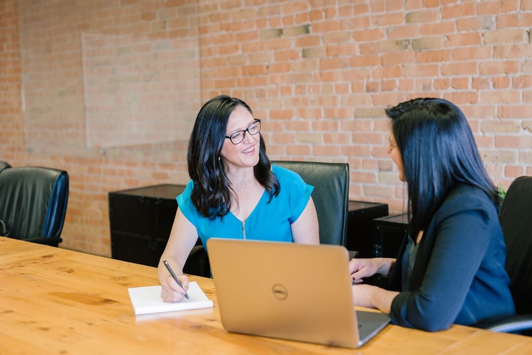 Two women engaged in discussion at a table, each using a laptop, focused on knowledge management software.
